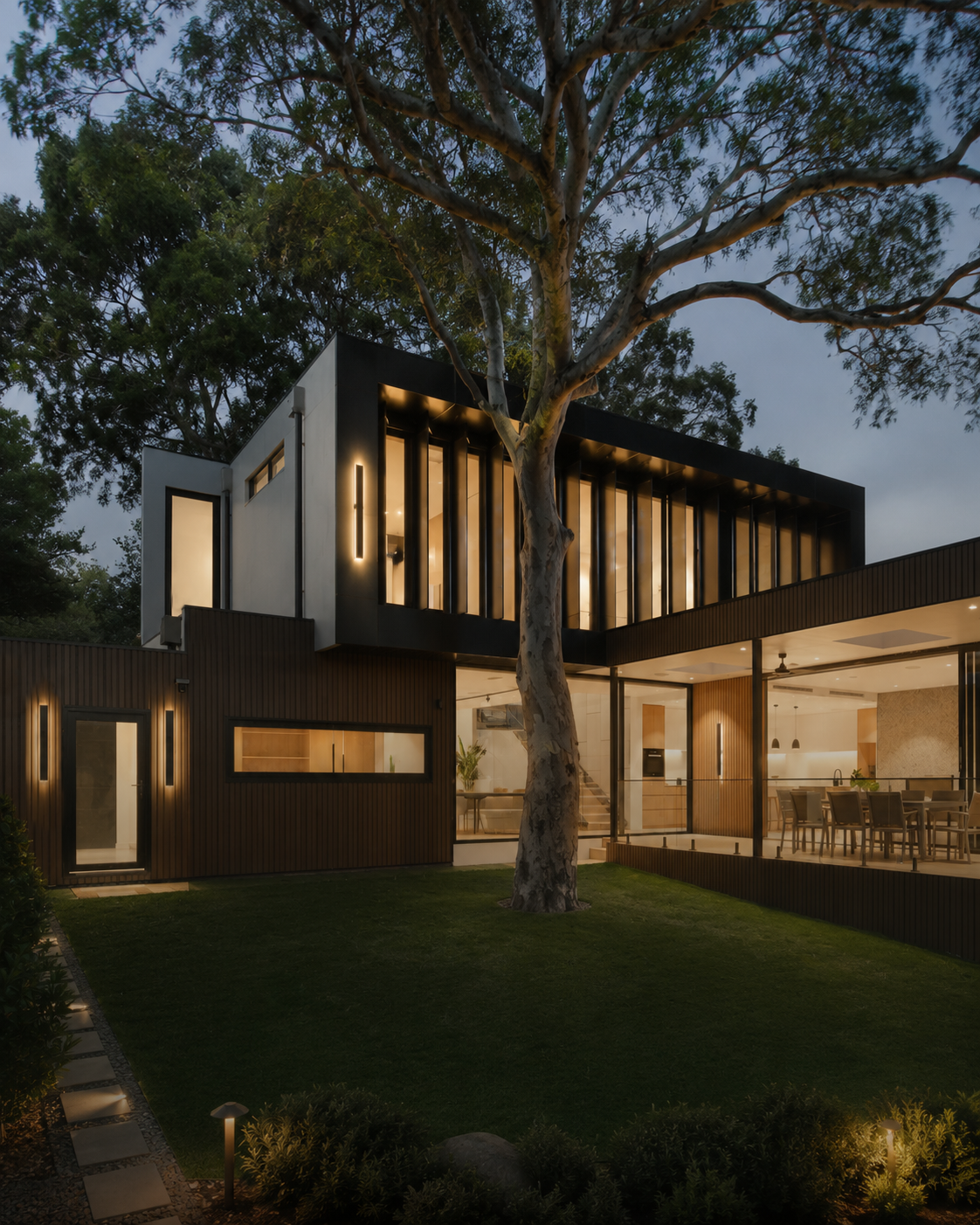 Modern two-story house exterior at dusk with dark wood and white paneling, large vertical window slats, warm interior lighting, a front lawn, landscaped shrubs, and a tall tree in the foreground.